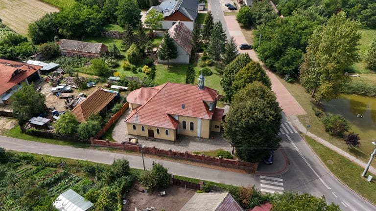 Fotografia lotnicza wsi Łuszkowo przedstawiająca kościół z dwuspadowym, czerwonym dachem i małą wieżyczką, otoczony ceglanym murkiem i zielenią. Wokół widoczna zabudowa wiejska, droga asfaltowa z przejściem dla pieszych, tereny zielone i fragment stawu.