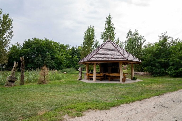 Zdjęcie przedstawia otwartą, drewnianą altanę znajdującą się przy stawie w Łuszkowie. Wewnątrz altany znajdują się drewniane ławki i stół. Altana stoi na zielonym trawniku, otoczona drzewami i krzewami
