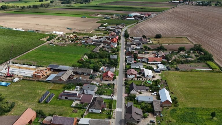 Fotografia lotnicza przedstawiająca wieś Łuszkowo z zabudową rozproszoną wzdłuż drogi, otoczoną polami uprawnymi o zróżnicowanej kolorystyce. Widoczne domy jednorodzinne z ogrodami, budynki gospodarcze, fragmenty lasów/zadrzewień oraz elementy infrastruktury wiejskiej.