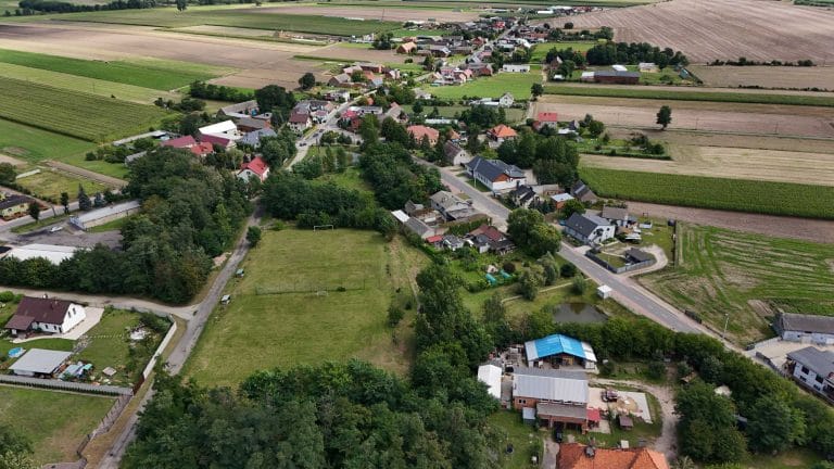 Fotografia lotnicza przedstawiająca wieś Łuszkowo. Widać zabudowę, z domami jednorodzinnymi i budynkami gospodarczymi, otoczoną polami uprawnymi. Widoczne drogi asfaltowe, drzewa i krzewy.