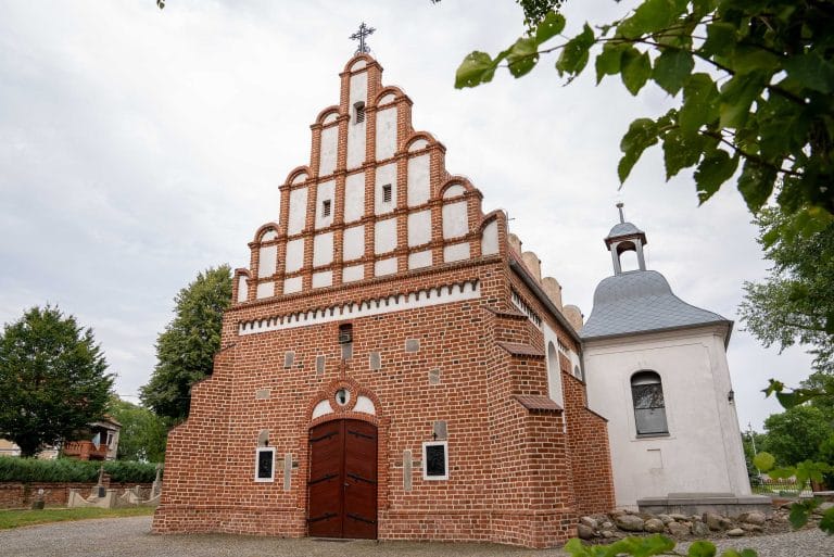 Zdjęcie przedstawia kościół rzymskokatolicki pw. św. Apostołów Piotra i Pawła w Rąbinie z charakterystycznym, schodkowym szczytem zdobionym białymi elementami. Do kościoła przylega biała, murowana wieżyczka z dzwonem, zwieńczona szarym, stożkowym dachem. Przed kościołem widoczny fragment zieleni.