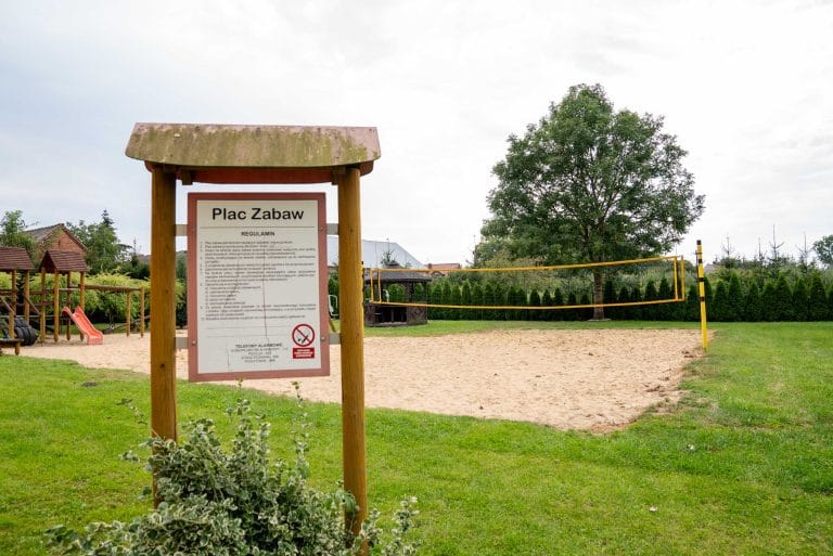 Zdjęcie przedstawia plac zabaw w Rąbinie. Na pierwszym planie widoczny jest regulamin placu zabaw, a za nim można zauważyć boisko do siatkówki. Po lewej stronie od boiska widać zjeżdżalnie i inne urządzenia. Za boiskiem widać altanę. Całość jest otoczona zielenią.