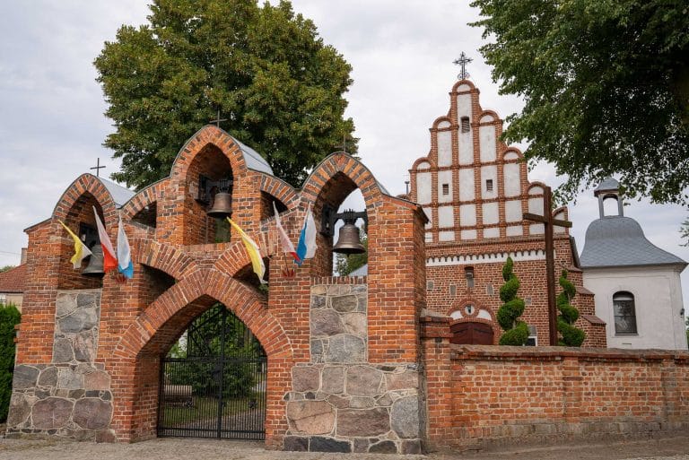 Zdjęcie przedstawia bramę przed kościołem rzymskokatolickim pw. św. Apostołów Piotra i Pawła w Rąbinie z trzema arkadami, w których wiszą dzwony. Na bramie powiewają flagi. W tle widoczny fragment kościoła i zieleni.
