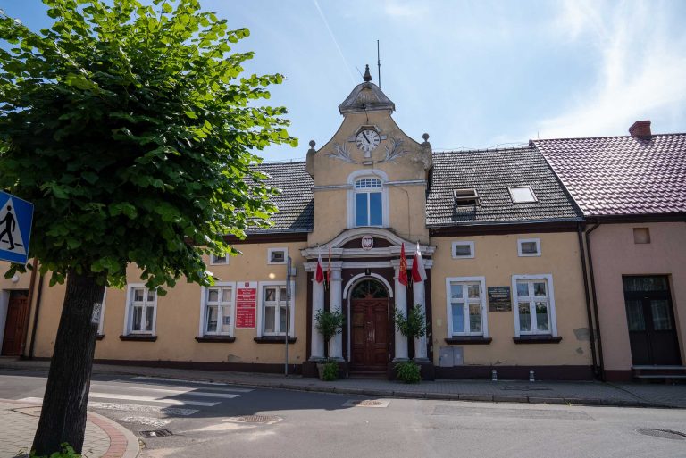 Na zdjęciu widoczny jest budynek Urzędu Miasta i Gminy Krzywiń. Jest to dwukondygnacyjny budynek o jasnej, beżowo-żółtej fasadzie, z ciemnym, dwuspadowym dachem pokrytym dachówką. Na centralnej części fasady umieszczony jest zegar. Nad wejściem głównym, do którego prowadzą schody, wisi godło oraz dwie flagi Polski. Okna budynku są prostokątne, z białymi ramami. Przed budynkiem, po lewej stronie, rośnie duże drzewo liściaste, częściowo zasłaniając widok.