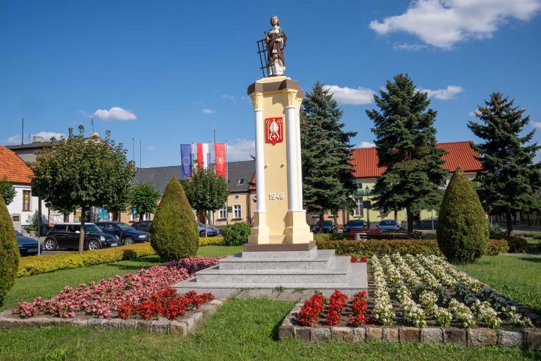 Na zdjęciu widoczny jest pomnik św. Wawrzyńca w Krzywiniu. Pomnik stoi na niewielkim placu otoczonym zielenią i niskimi drzewami. Składa się z jasnego, kamiennego cokołu, na którym umieszczona jest figura świętego. Na cokole widnieje data "1946". Na przedniej ścianie cokołu umieszczony jest herb Krzywinia.