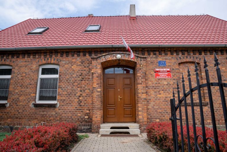 Zdjęcie przedstawia budynek świetlicy wiejskiej w Wieszkowie. Zbudowany jest z czerwonej cegły, ma dwuspadowy dach pokrytym czerwoną dachówką. Nad wejściem wisi polska flaga. Przed budynkiem rosną krzewy.