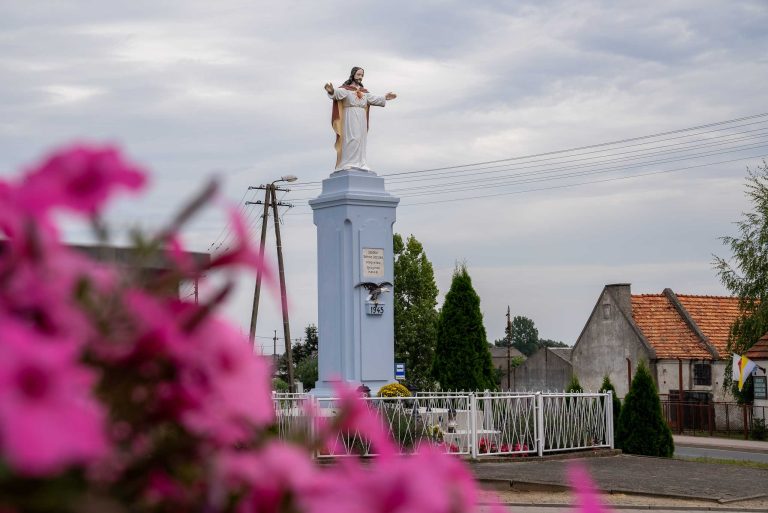 Zdjęcie przedstawia figurę Chrystusa Króla w Wieszkowie z rozłożonymi rękami, stojącą na wysokim, jasnym postumencie. Figura otoczona jest niskim ogrodzeniem. W tle widać zabudowania wiejskie.