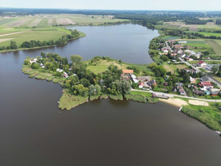 Fotografia lotnicza przedstawia rozległe jezioro przy wsi Zbęchy. Wzdłuż brzegu widoczna jest zabudowa wiejska i pola uprawne. W oddali rozciągają się lasy i pagórkowaty krajobraz.