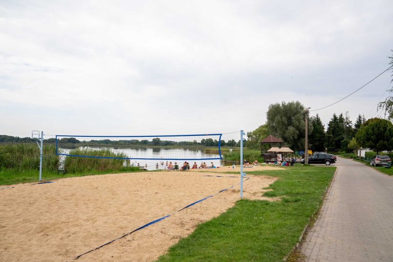 Zdjęcie przedstawia piaszczyste boisko do siatkówki plażowej z rozciągniętą siatką przy jeziorze Zbęchy. W tle rozległe jezioro, a na brzegu siedzących i odpoczywających ludzi. Obok boiska znajduje się droga, zaparkowane samochody i dużo zieleni.