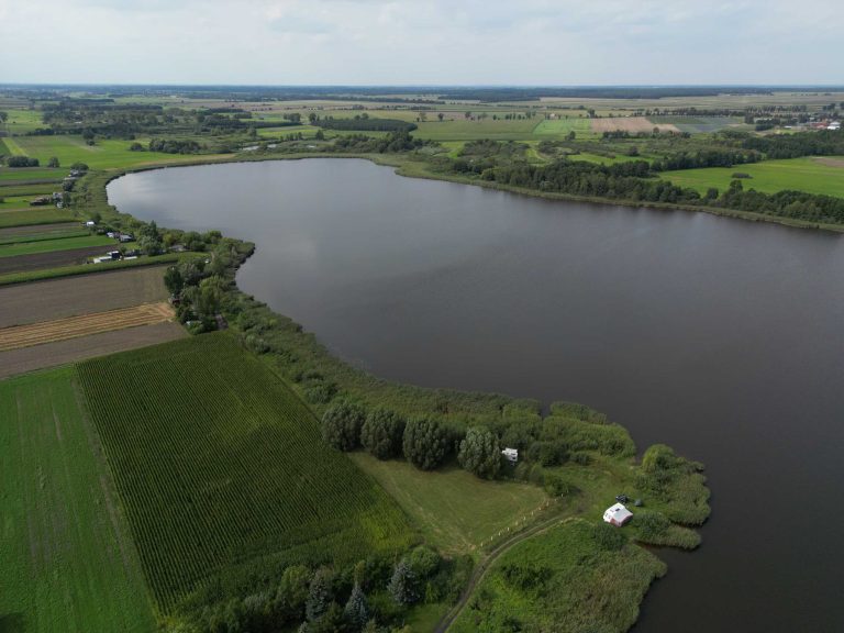 Fotografia lotnicza przedstawia rozległe jezioro Zbęchy o nieregularnym kształcie, otoczone polami uprawnymi i zielenią. W oddali widać lasy i zabudowania.