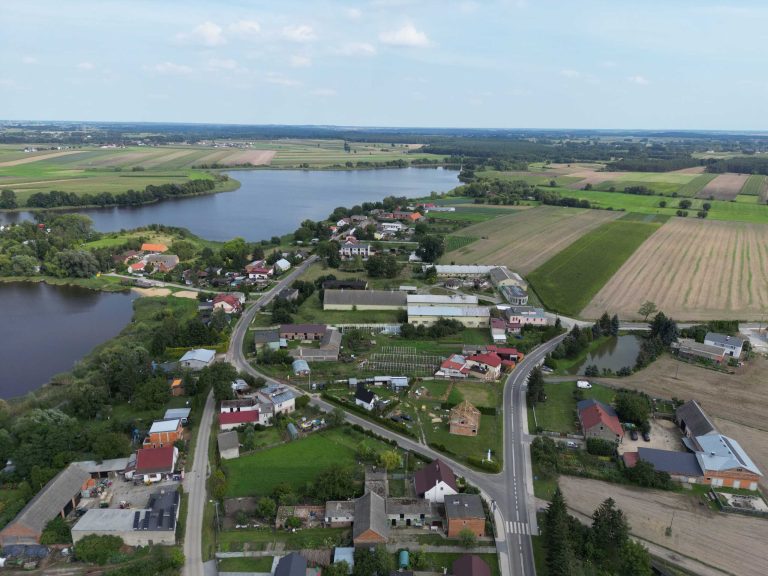 Fotografia lotnicza przedstawia wieś Zbęchy która jest położona w pobliżu jeziora. Widoczne są domy, drogi, pola uprawne i zbiornik wodny. W oddali widać lasy.