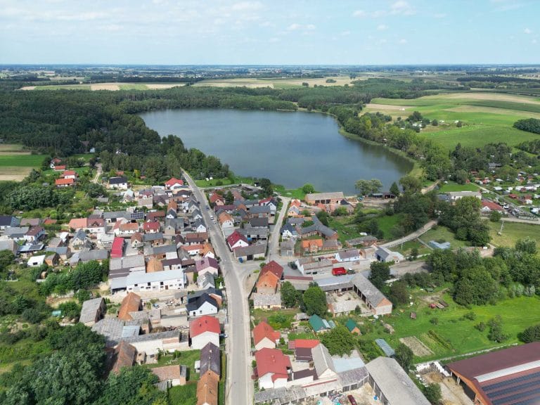 Fotografia lotnicza przedstawiająca wieś Żelazno oraz jej jezioro. Widać gęstą zabudowę z licznymi domami i gospodarstwami. Wokół jeziora można zauważyć liczną zieleń, lasy. Na horyzoncie widać pola uprawne.