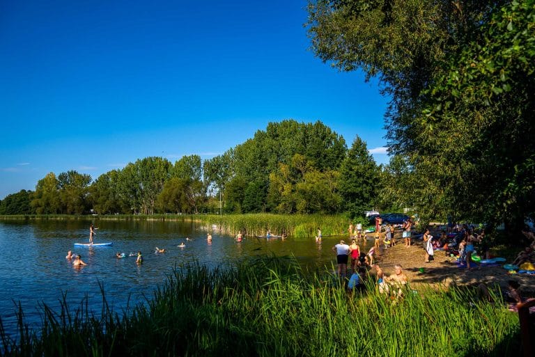Zdjęcia przedstawia plażę przy jeziorze Żelazno. Widac na niej dużą ilość osób które odpoczywają lub pływają. Na wodzie można zauważyć łódkę. Całość jest otoczoan bujną zielenią.
