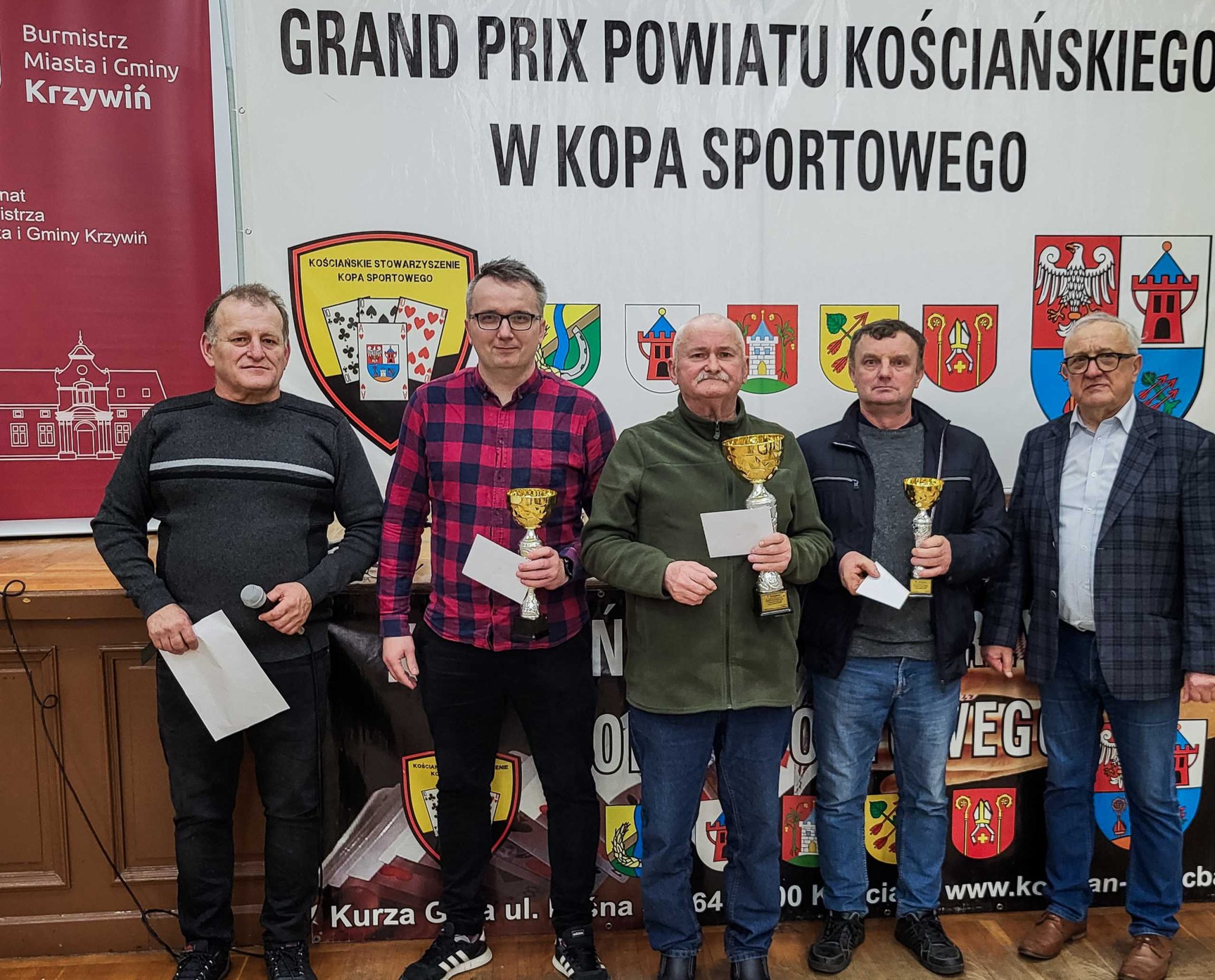 Pięciu mężczyzn stoi na tle baneru z napisem 'Grand Prix Powiatu Kościańskiego w Kopa Sportowego'. Trzech z nich trzyma złote puchary i koperty, co sugeruje nagrody za osiągnięcia w turnieju. Mężczyzna po lewej trzyma mikrofon i kartkę, a ostatni z prawej nosi okulary i elegancką marynarkę. W tle widać herby gmin oraz logo Kościańskiego Stowarzyszenia Kopa Sportowego. Wydarzenie odbywa się pod patronatem burmistrza Miasta i Gminy Krzywiń.