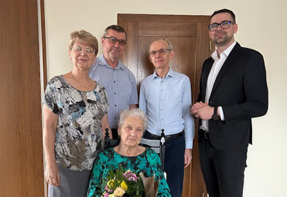 Grupa pięciu osób pozuje do zdjęcia w pomieszczeniu. Starsza kobieta z bukietem kwiatów siedzi na krześle, otoczona przez trzy osoby w średnim wieku i jednego młodszego mężczyznę w garniturze. Wszyscy patrzą w obiektyw i uśmiechają się.