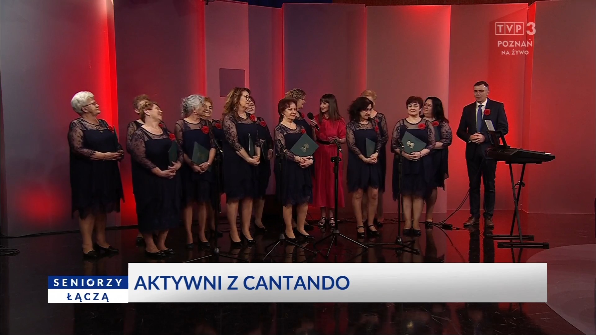 Grupa seniorek z chóru Cantando w granatowych sukienkach z koronkowymi rękawami i czerwonymi różami przypiętymi do stroju, stoi na scenie telewizyjnej TVP3 Poznań. Obok nich znajduje się prowadząca w czerwonej sukni oraz mężczyzna w garniturze przy keyboardzie. W tle czerwone oświetlenie oraz napis 'Seniorzy łączą – Aktywni z Cantando'.