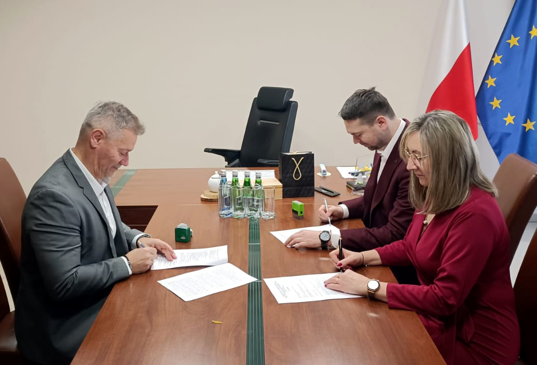 Zdjęcie prezentujące podpisanie umowy. Trzy osoby, w tym Burmistrz, siedzą przy stole i podpisują dokumenty. W tle widać flagę Polski i UE.