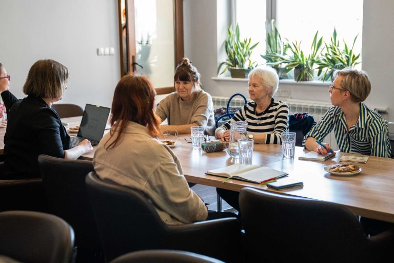 Grupa 5 kobiet siedzi przy stole i rozmawia. Znajdują się w pomieszczeniu z białymi ścianami. Na wspomnianych stołach stoi dzbanek z wodą oraz talerzyk z ciasteczkami, leżą też notatniki i dokumenty.