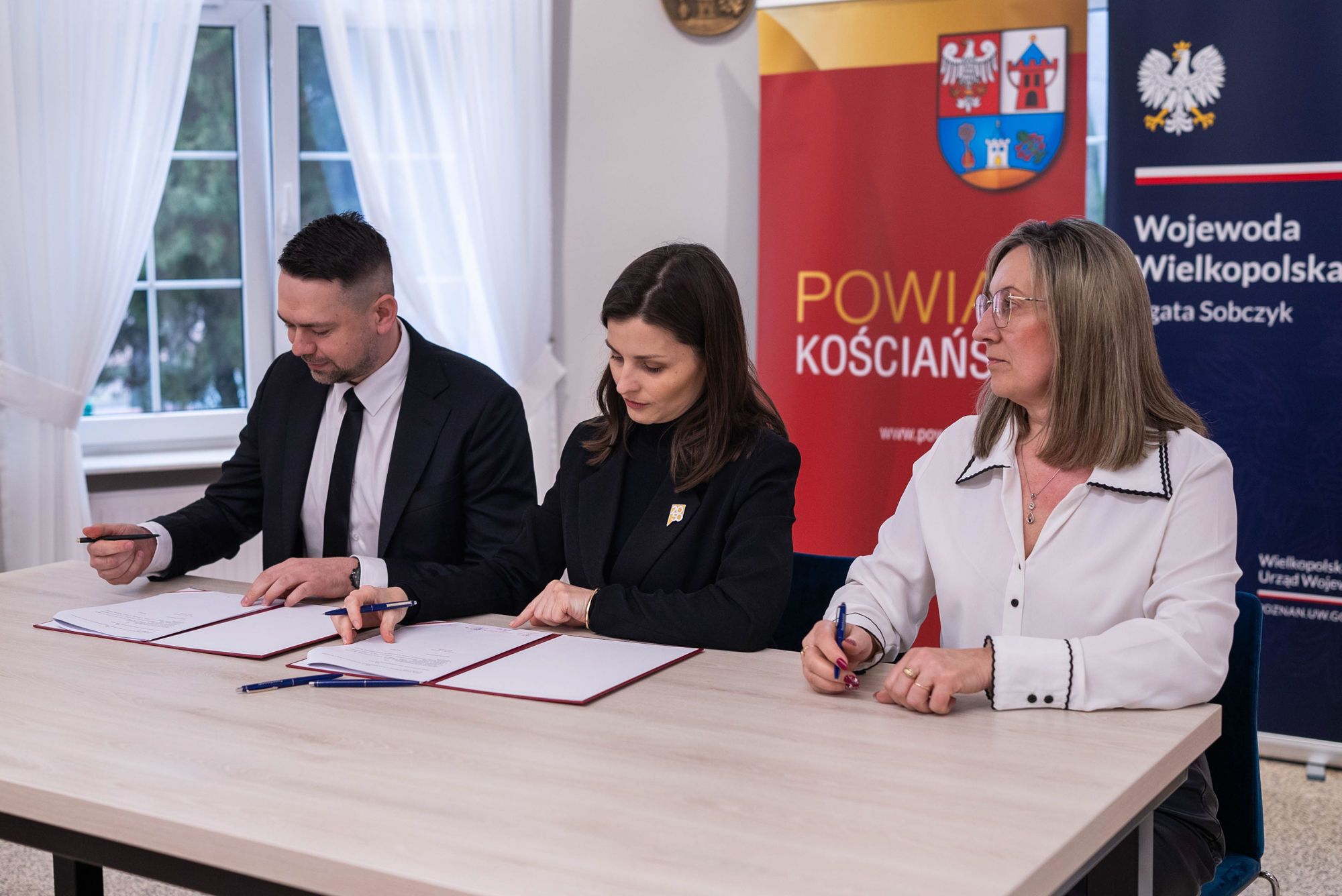 Trzy elegancko ubrane osoby podpisują dokumenty siedząc obok siebie przy stole.