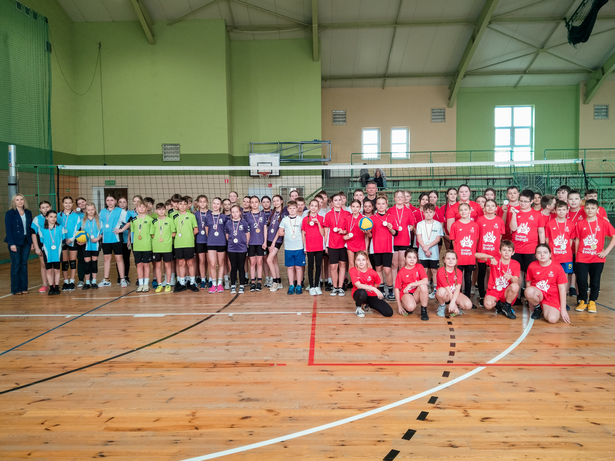 Grupa dzieci i młodzieży w kolorowych strojach sportowych (niebieskich, zielonych, fioletowych i czerwonych) pozuje w hali sportowej przed siatką do siatkówki. Wszyscy mają na szyjach medale, a niektóre osoby trzymają piłki do siatkówki. Obok stoi dorosła kobieta w eleganckim stroju. W tle widać trybuny i zielone ściany hali.