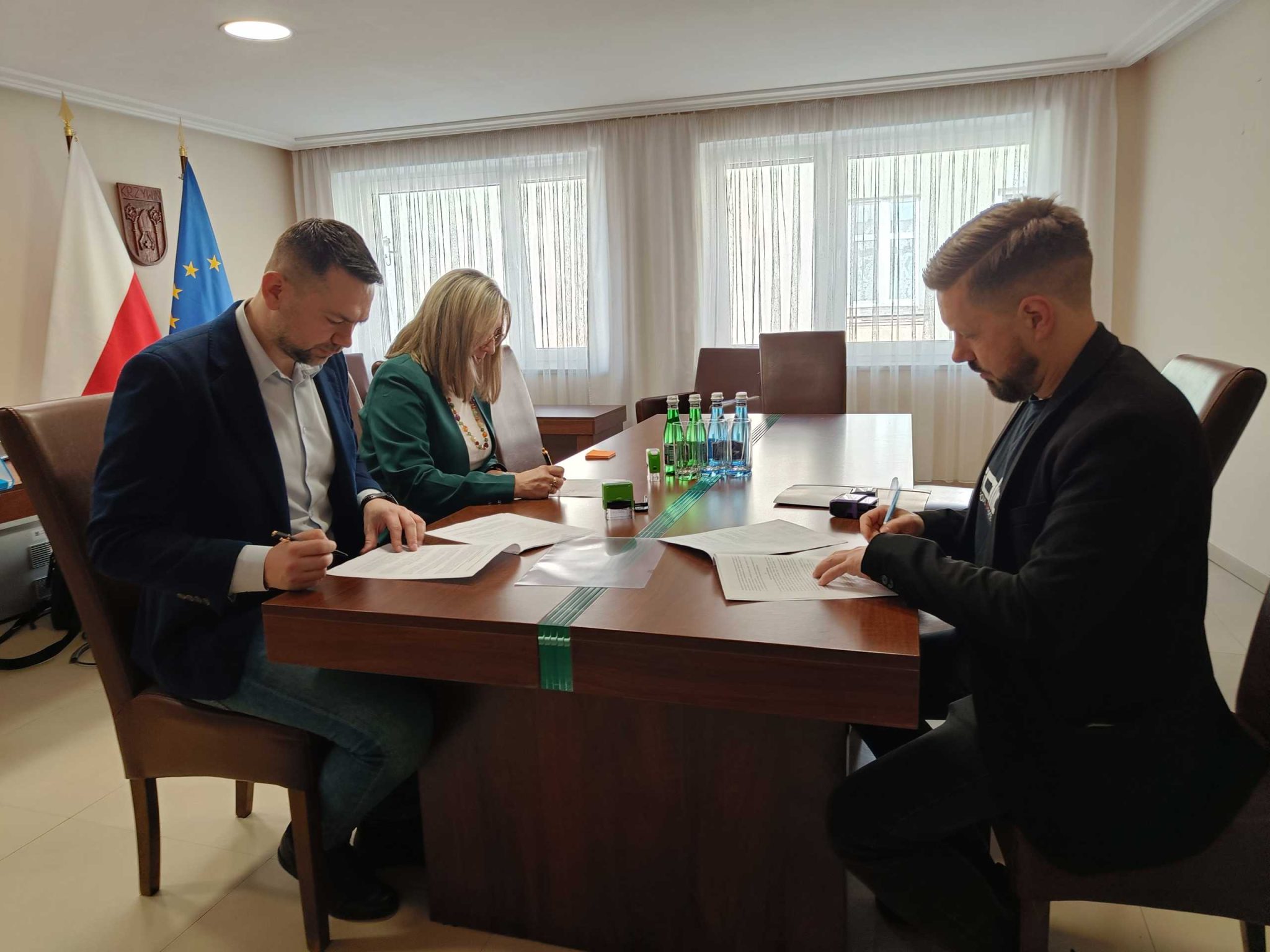 Fotografia przedstawia trzy osoby ubrane w elegancką odzież, siedzące przy stole w gabinecie, w tle widać flagę Polski oraz UE. Osoby przy stole podpisują dokumenty.