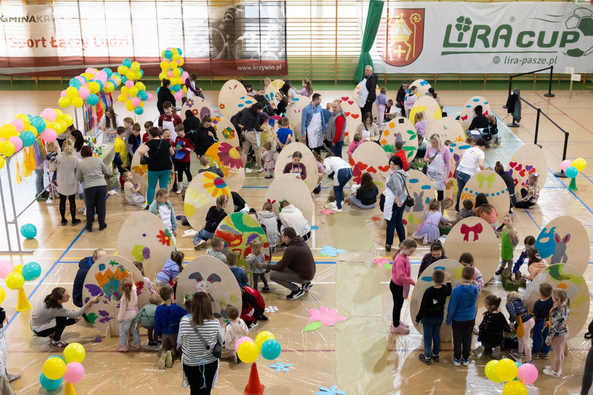Dzieci i dorośli uczestniczą w wielkanocnych warsztatach plastycznych w hali sportowej, wspólnie malując duże, tekturowe pisanki. W tle kolorowe balony, dekoracje i banery reklamowe. Wydarzenie odbywa się w przyjaznej, rodzinnej atmosferze.