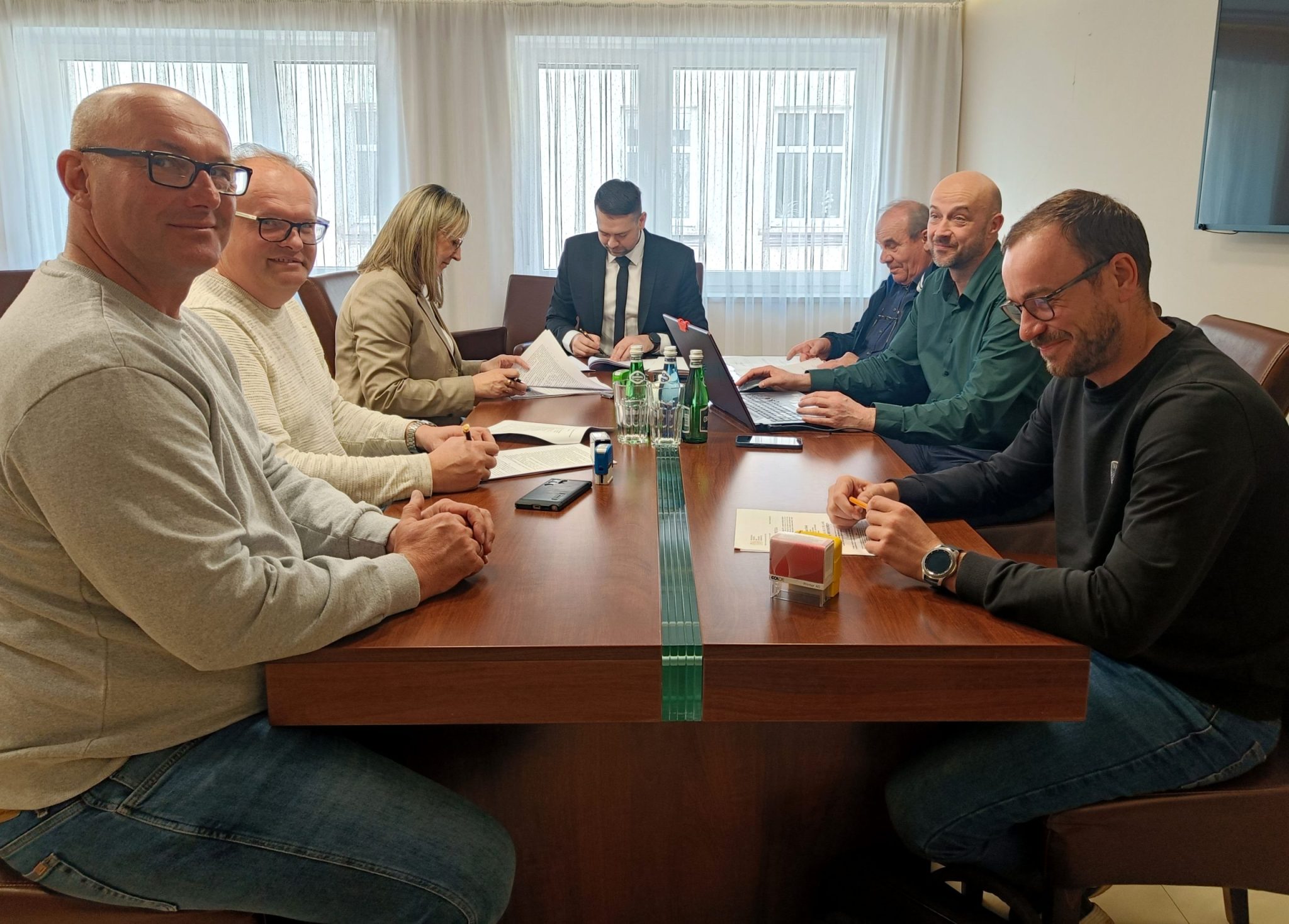 Grupa osób zaangazowanych w projekt siedzi przy urzędowym stole i podpisuje umowy