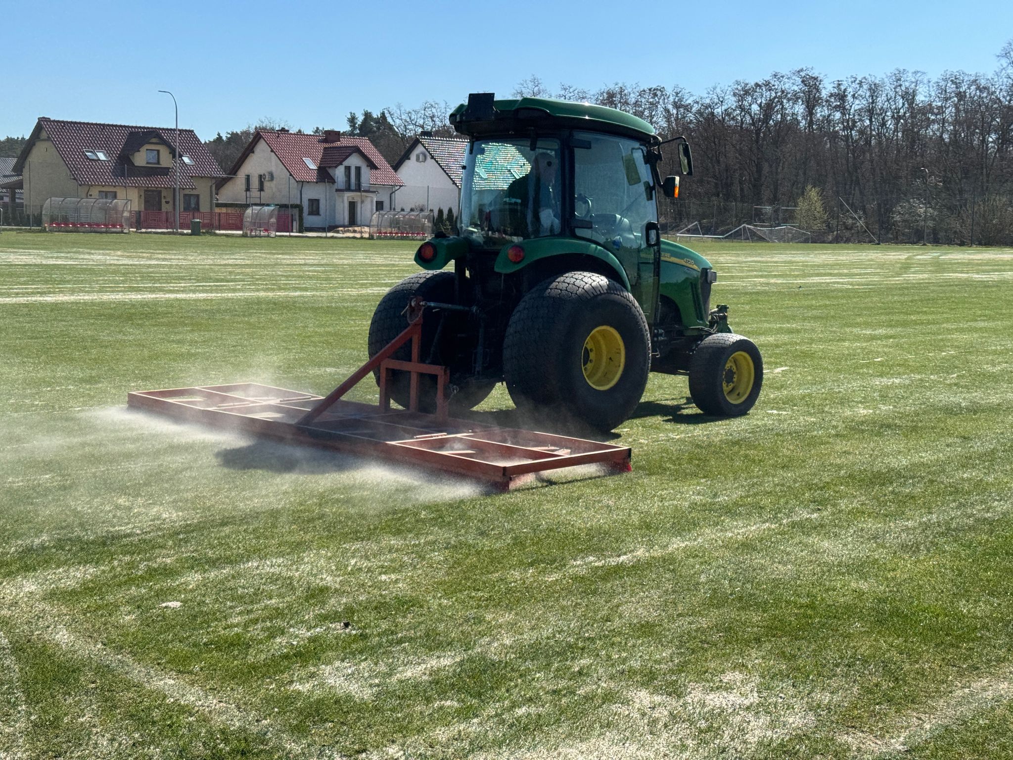 Traktor jadący po murawie boiska, ciągnący przyrząd służący do pielęgnacji trawy