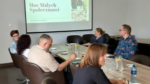 Fotografia przedstawia osoby siedzące przy stole i uczestniczące w spotkaniu. Patrzą na ekran projektora.