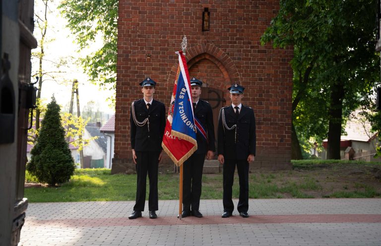 Trzech młodych strażaków w mundurach galowych stoi na tle budynku i parku i trzyma sztandar.