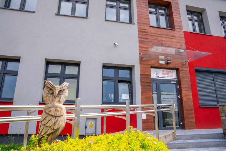 Zdjęcie przedstawia budynek biblioteki z zewnątrz, z widoczną nazwą, rampą dla niepełnosprawnych oraz sową wyrzeźbioną z drewna