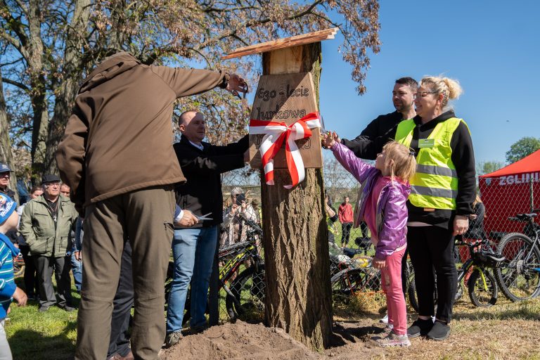 Przedstawiciele odsłaniają drewnianą tablicę przyczepioną do drzewa, z napisem 630-lecie Jurkowa