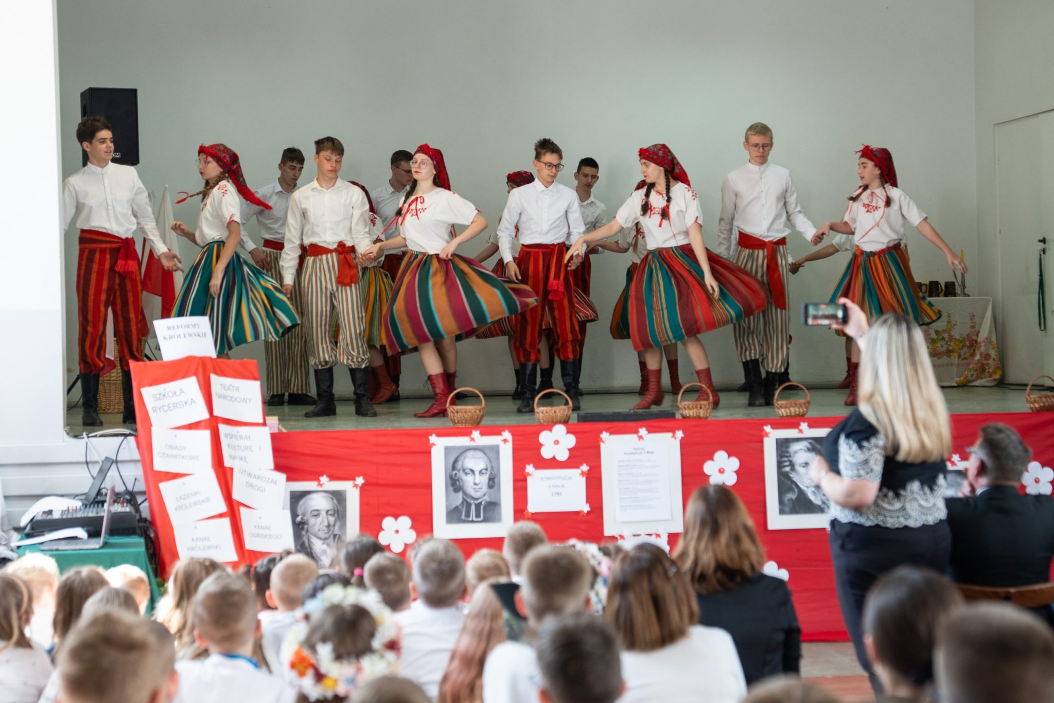 Dzieci występują na scenie podczas gdy inna grupa uczniów obserwuje występ. Uroczystość z okazji święta III Maja.
