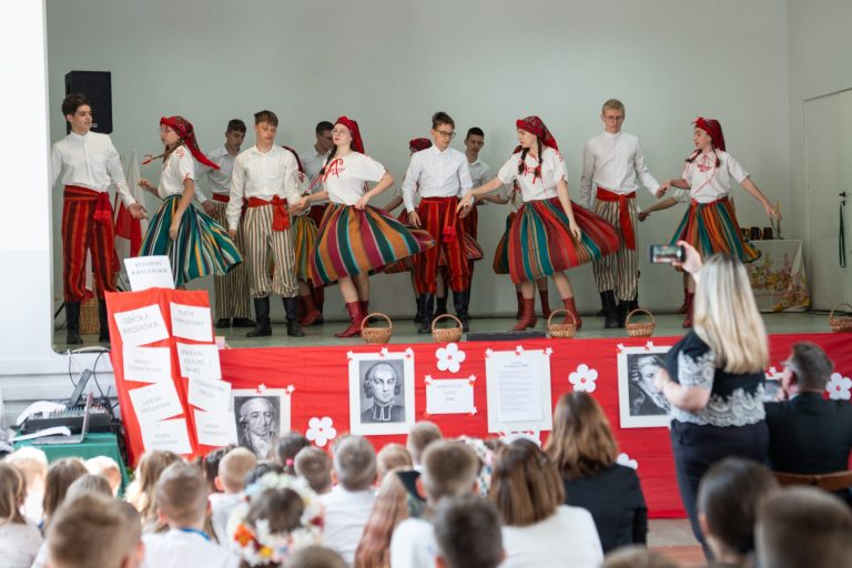Dzieci występują na scenie podczas gdy inna grupa uczniów obserwuje występ. Uroczystość z okazji święta III Maja.