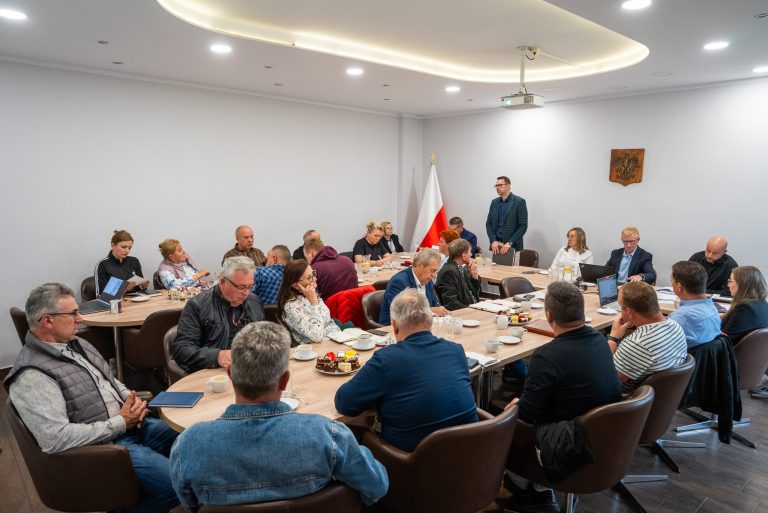 Spotkanie w sali konferencyjnej z udziałem kilkudziesięciu osób siedzących przy stołach. Jeden z mężczyzn stoi i przemawia. Na ścianie widoczna flaga Polski oraz godło. Uczestnicy mają przed sobą dokumenty, laptopy i poczęstunek. Atmosfera formalna.