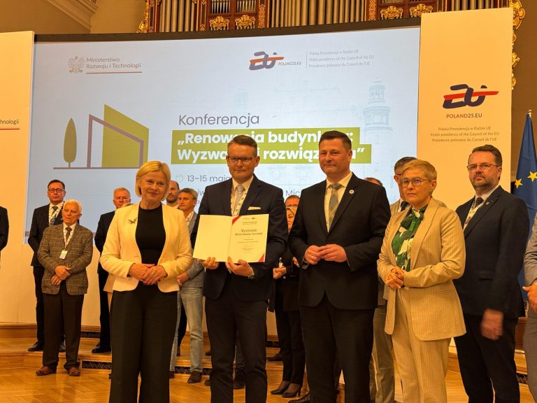 Grupa elegancko ubranych osób stoi w sali na tle ekranu; jedna z osób trzyma w rękach dokument - wyróżnienie