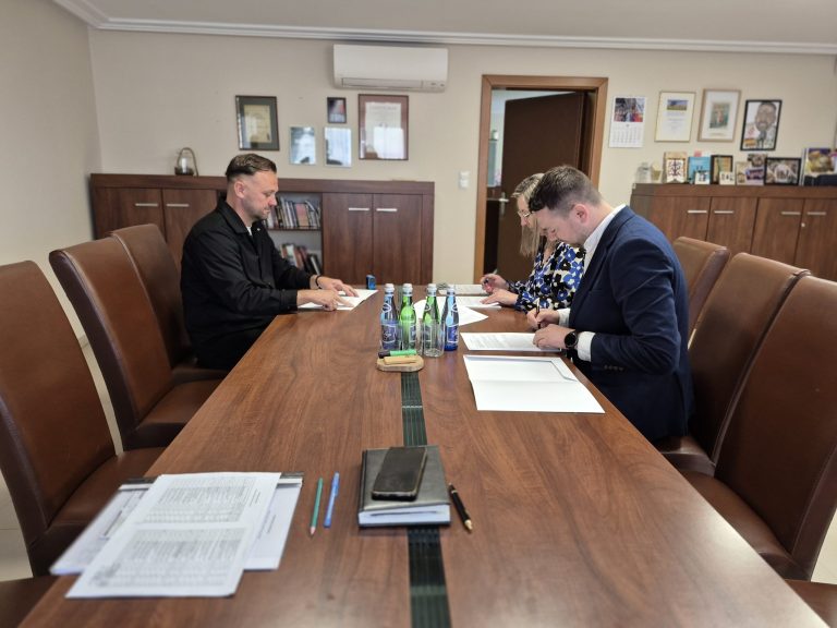 Moment podpisania umowy w gabinecie burmistrza w Urzędzie Miasta i Gminy. Dwie elegancko ubrane osoby siedzą naprzeciw siebie, na stole leżą dokumenty.
