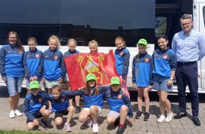 Fotografia przedstawia młode zawodniczki w sportowych dresach klubowych z trenerem i flagą na tle autobusu