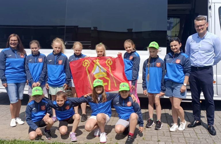 Fotografia przedstawia młode zawodniczki w sportowych dresach klubowych z trenerem i flagą na tle autobusu