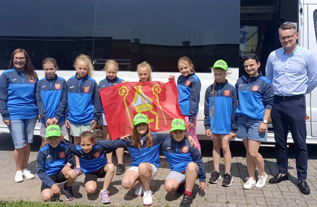 Fotografia przedstawia młode zawodniczki w sportowych dresach klubowych z trenerem i flagą na tle autobusu